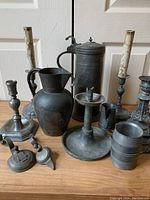 Full group shot of all pewter items arranged on wooden floor showing pitcher, jug, multiple candle holders, snuffers, and small containers.