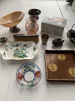 Assorted ladies dresser accessories laid out on table including trays, small vases, brush, box, and metal basket.