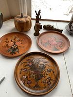 View of three handpainted wooden wall plates with floral decoration, carved wooden pumpkin, carved wooden bust, and two other carved wood items on a white surface by window.