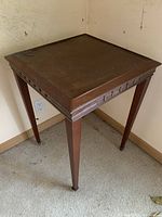 Full view of the small square wooden side table against the wall showing the top surface and legs.