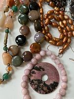 Close-up of various bead necklaces including round polished rose quartz, mixed stone beads, and copper-toned freshwater pearls.