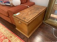 Antique hardwood blanket box photographed indoors beside an orange couch and wooden floor, showing worn top with scratches and cracks and brass drawer pull on the side.