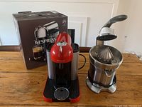 Nespresso Vertuo coffee maker in red with box and Breville automatic citrus press stainless steel model 800CPXL on wooden table.