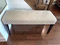 Front view of wooden frame bench with beige geometric patterned upholstered padded seat, standing on hardwood floor against white wall.