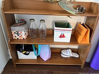 Wood bookcase with three shelves and various items on shelves including two glass bottles, a white box, slippers, and other miscellaneous items. Wood has a medium finish with natural grain showing.