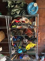 Shelving unit with folded and rolled green tarp, blue water jug, various ropes, straps, and backpacks stacked on metal shelves.
