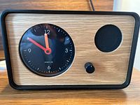 Front view of oneclock alarm clock showing wood finish front panel, analog clock face with orange and red hands, clock brand, black frame, control knob and speaker grill.