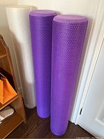 Three foam rollers standing upright: two purple textured rollers and one smooth white roller.