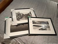 Photo showing three framed vintage black and white photo prints stacked on a bedspread with corner protectors on two frames.