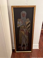 Full view of a tall rectangular framed brass rubbing showing a knight in armor holding a shield and sword, gold frame, black background.