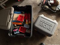 Black plastic bin containing assorted hardware, small boxes, red plastic containers, sandpaper, and other miscellaneous items, open and showing contents.