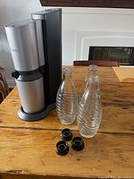 Silver SodaStream sparkling water maker with black base, 3 spiral glass bottles and 3 black caps on wooden table