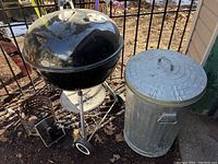 Photo of black Weber kettle charcoal BBQ grill with silver legs, wheels, and Oklahoma chimney charcoal starter next to metal garbage can with lid.