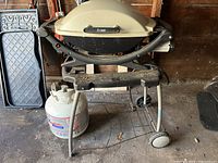 Front view of Weber Q200 BBQ on metal stand with propane tank placed beside it in a garage setting.