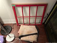 Antique painted wooden window pane with red frame and eight glass panes resting against the wall with fabric wall hanging folded below it.