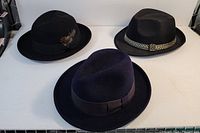 Three fedora hats on white surface, two black vintage with feather detail and one newer navy blue without feathers