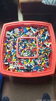 Top-down view of red Lego table filled with assorted Lego pieces in multiple colors and shapes, showing the volume and variety of pieces.