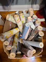 Photo of box containing around 23 rolls of vintage and mid-century modern wallpaper in floral and geometric patterns. Some rolls sealed, some opened.