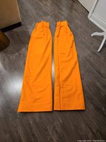 Two orange drapes laid flat side by side on wood flooring showing pleated tops and clean edges.