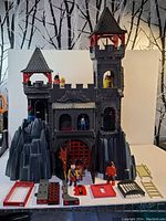Front view of the Playmobil dragon castle with figures on the battlements and gate, showing rock side structures and several castle parts on the ground.
