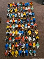 Top view showing all 89 Playmobil figures arranged in rows on a carpeted floor, displaying a wide range of colorful clothing and headgear.