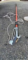 Tree pruner pole close-up showing compound pulley mechanism, cutting arm, and attached white rope on asphalt surface.