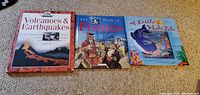 Three large hardcover storytime books lay flat on carpet: 'Volcanoes & Earthquakes', 'The Best Book of Pirates', and 'A Little Whale Tale'. Covers show colorful illustrated designs corresponding to each book's theme.