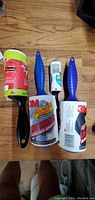 Photo showing four lint rollers laid on wooden floor, including two 3M lint rollers with blue handles, one small blue-handled roller, and one Scotch-Brite Sticky Sheets container.