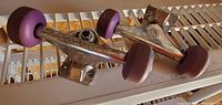 Two ML skateboard trucks each fitted with four purple wheels laid on a white surface, showing the side profile of both.