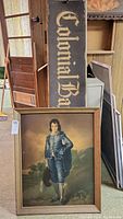 Framed portrait of a boy in blue attire and vintage wooden Colonial sign leaning against each other in a storage room.
