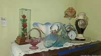 Photo of vintage glass baskets, clock lamp, alarm clock, decorative vases and glass tray arranged on a wooden surface with a lace cloth.
