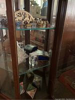 View of glass shelves inside a wooden display cabinet showing ceramic figurines including animals, paperweight, decorative box, shell and paper ornaments