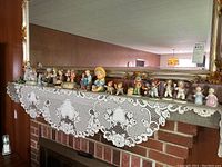Wide shot of all figurines arranged on a mantel over lace cloth showing entire collection of various small collectibles including angels, children, and animal figurines.