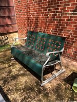 Full view of green metal patio glider bench with curved silver arms and legs, placed outdoors against a brick wall.