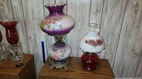 Two vintage lamps on wooden furniture against wood panel wall; a 21-inch Gone with the Wind style electric lamp with pink floral painted glass and ornate metal base, and a 16-inch oil lamp with red glass base and floral painted milk glass shade.