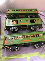 Three Lionel train cars: two Pullman passenger cars and one mail car, green with orange accents, arranged side by side on a cloth surface.
