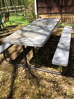 Full view of rectangular picnic table with attached bench seats, white plastic top and seats on dark metal frame, outdoors on grass and dirt with leaf debris.