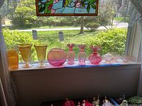 Photo of various colored glass pieces placed on a window ledge with natural sunlight showing details of texture and color including yellow vases, pink glass vases, bowls, and an orange container.