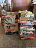Photo showing a stack of vintage board games and model kits including Mayflower, Dealer's Choice, Hollywood Squares, NFL VCR Quarterback, Masterpiece, Risk, and a Canadian Mist box used for storage.