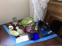 Photo of vintage glass figurines on a wooden surface including various birds, a green turtle, an elephant, a swan, and a blue multifaceted paperweight.