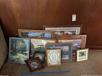 Multiple framed prints leaning against a wood-paneled wall showing vintage cars, trains, and winter landscape scenes.