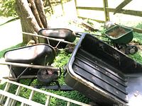 Two black metal wheelbarrows outdoors, visible metal trays and handles, some rust and dirt, one tire appears flat.