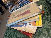 Framed fashion prints and vintage magazines stacked on a green patterned fabric surface, shows wear and condition of items.