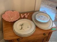 View of all ceramic items on wooden chest, showing plates stacked with green leaf border and palm tree design, pink leaf bowl, and pink salt and pepper bird figurines.