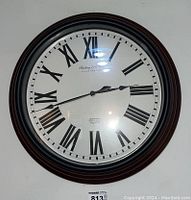 Front view of the Sterling and Noble wall clock showing the full clock face with Roman numerals and black hands on a white dial under glass. The metallic frame has a dark finish with reddish and black layers.