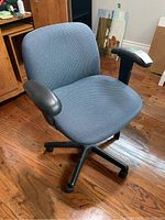 Blue fabric upholstered desk chair with black adjustable armrests and black caster base, placed on wooden floor.