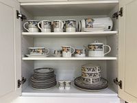 Three shelves of white ceramic Mikasa dishes with colorful floral and fruit pattern including mugs, plates, and serving items.