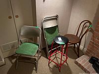 Four folding metal chairs with green vinyl seats and backrests, metal folding frame in beige. Also a wooden chair with curved backrest and a metal stool with red frame and black vinyl round seat. Chairs and stool show some wear.