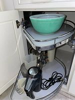 Under sink pull-out shelf showing green ceramic mixing bowl and Black & Decker hand mixer with beaters and power cord.