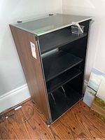 Side angle view showing the wood-grain laminate sides and clear glass top with protective corner pads. The cabinet sits on caster wheels and is empty.